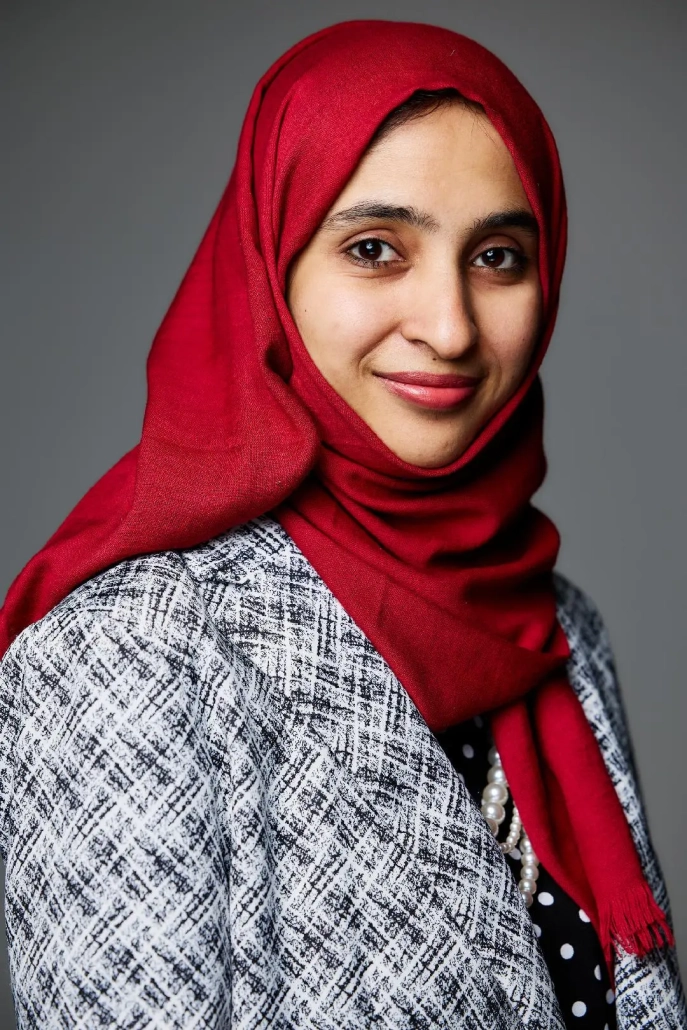 Dr Faiza Ali in a red hijab and patterned blazer smiles softly against a gray background.