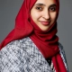 Dr Faiza Ali in a red hijab and patterned blazer smiles softly against a gray background.