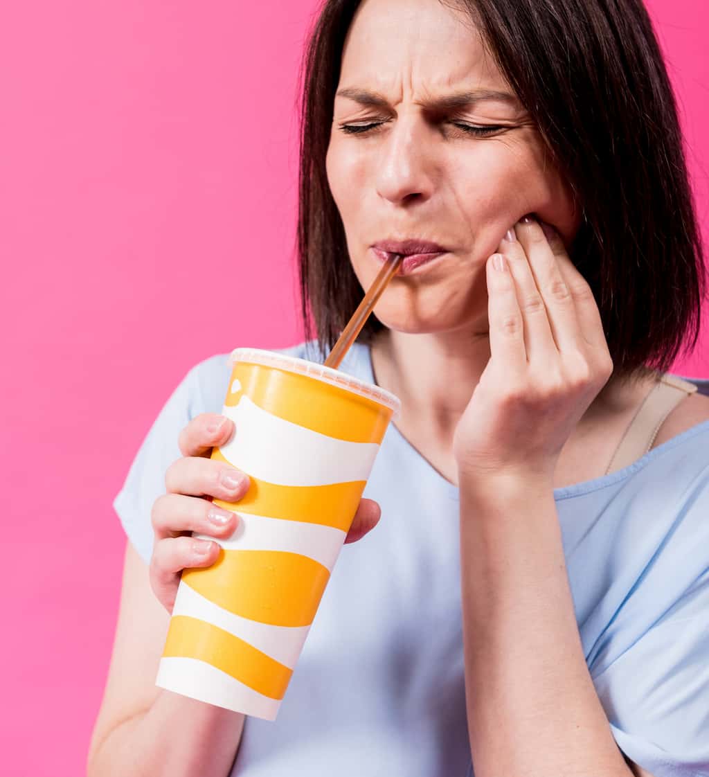 Woman drinking from a cup while holding her cheek in discomfort against a pink background.