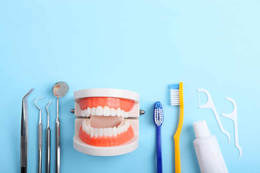 Dental tools and hygiene products, including a toothbrush and toothpaste, are arranged around a model of teeth on a blue background.