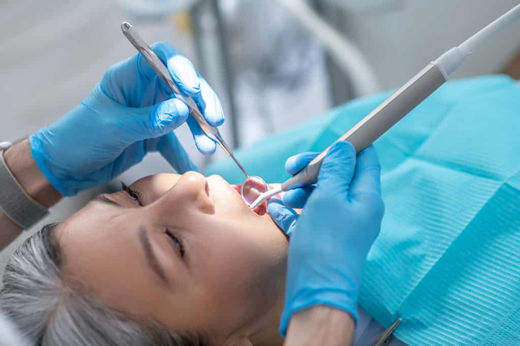 Dentist examines a patient's mouth using dental tools, wearing blue gloves.