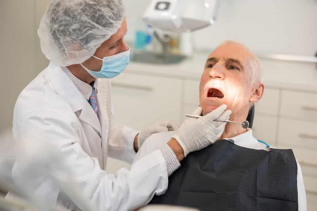 A dentist examines an elderly patient's mouth under a bright light in a dental clinic.